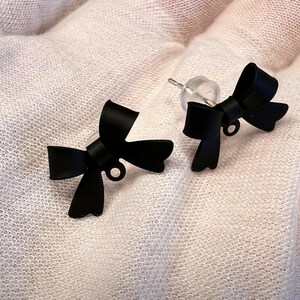 Könnte beinhalten: Ein Paar schwarze Schleifenohrringe. Die Schleifen bestehen aus einem glatten, glänzenden Material und haben ein schlichtes, elegantes Design.