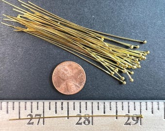 Può includere: Una collezione di spilli dorati con una piccola sfera all'estremità, sparsi su una superficie scura. Sono presenti anche una moneta da un centesimo e un righello, con uno spillo lungo circa 2,5 cm.