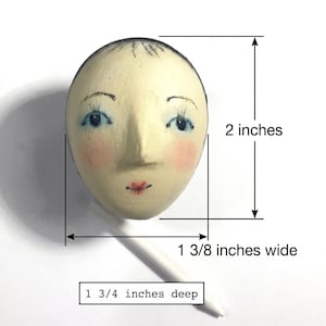 May include: A hand-painted wooden egg-shaped face with blue eyes, pink cheeks, and red lips. The object measures 2 inches in height, 1 3/8 inches in width, and 1 3/4 inches in depth, as indicated by the visible measurement labels.
