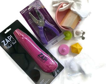 Può includere: Un utensile termico Zap! rosa nella sua confezione, una perforatrice McGill viola nella sua confezione, una ciotola rosa contenente un blocco di legno e tessuto bianco, un disco verde, un disco rosa, un oggetto giallo a forma di stella e ganci metallici.
