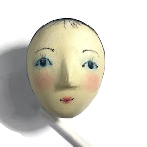 Pu&ograve; includere: Un oggetto di forma ovale color crema con un volto dipinto a mano, capelli scuri, occhi blu, guance rosa e labbra rosse. La superficie ha un aspetto strutturato e un sottile bastoncino bianco sporge dalla parte inferiore dell'oggetto.