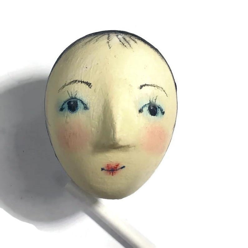 Pu&ograve; includere: Un oggetto di forma ovale color crema con un volto dipinto a mano, capelli scuri, occhi blu, guance rosa e labbra rosse. La superficie ha un aspetto strutturato e un sottile bastoncino bianco sporge dalla parte inferiore dell'oggetto.