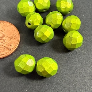 May include: A collection of nine small, round, faceted lime green beads with a metallic sheen. The beads are arranged in a cluster next to a copper-colored United States penny for scale, showing their relative size in relation to the coin.