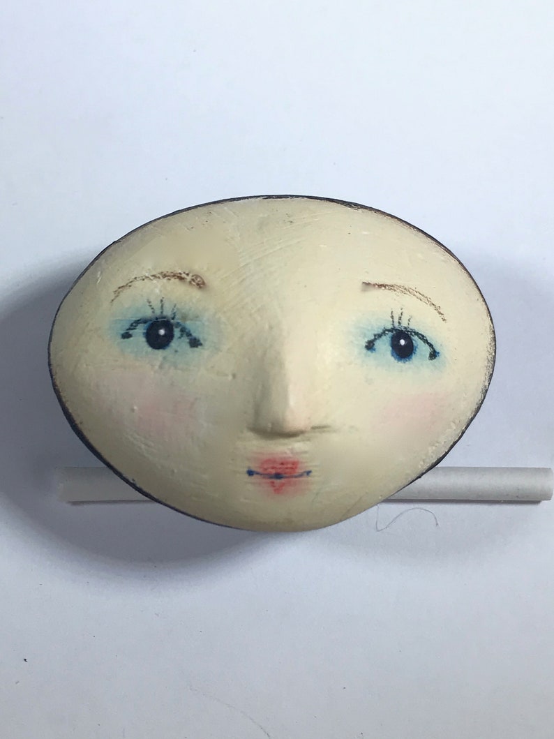 Peut inclure: Un visage en c&eacute;ramique peint &agrave; la main avec des yeux bleus, une bouche rose et un contour brun. Le visage est blanc avec une surface l&eacute;g&egrave;rement textur&eacute;e.