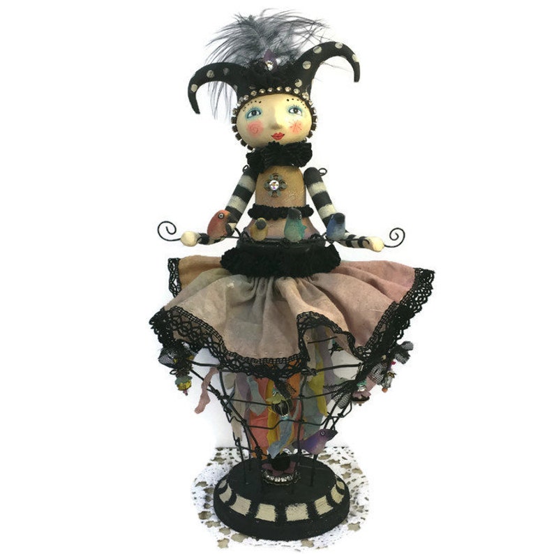 Peut inclure: Une poup&eacute;e fantaisiste avec une robe &agrave; rayures noires et blanches et un chapeau de jester, tenant une cage &agrave; oiseaux remplie d'oiseaux color&eacute;s. La poup&eacute;e est debout sur une base noire et blanche.