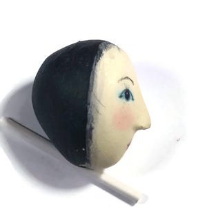Pu&ograve; includere: Una testa di bambola in legno dipinta a mano con un viso liscio color crema, tratti delicati, occhi azzurri, ciglia scure e guance rosa. La testa presenta un'acconciatura dipinta in nero solido ed &egrave; montata su un sottile bastoncino bianco.