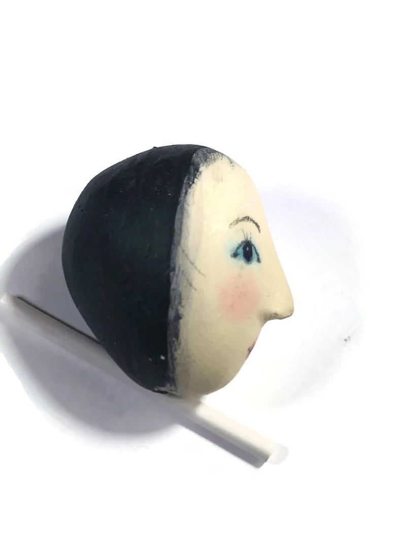 Pu&ograve; includere: Una testa di bambola in legno dipinta a mano con un viso liscio color crema, tratti delicati, occhi azzurri, ciglia scure e guance rosa. La testa presenta un'acconciatura dipinta in nero solido ed &egrave; montata su un sottile bastoncino bianco.