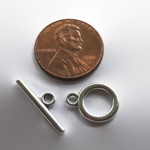 May include: Silver tone toggle clasp with a round ring and a bar. The clasp is shown next to a US penny for size comparison.