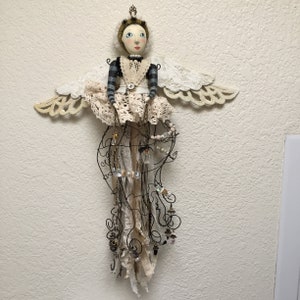 Pu&ograve; includere: Una bambola decorativa da parete con viso dipinto, corona dorata e ali in pizzo bianco. La bambola indossa una maglia a maniche lunghe a righe nere e grigie con un grembiule in pizzo bianco e una gonna fatta di fili, strisce di tessuto e perline.