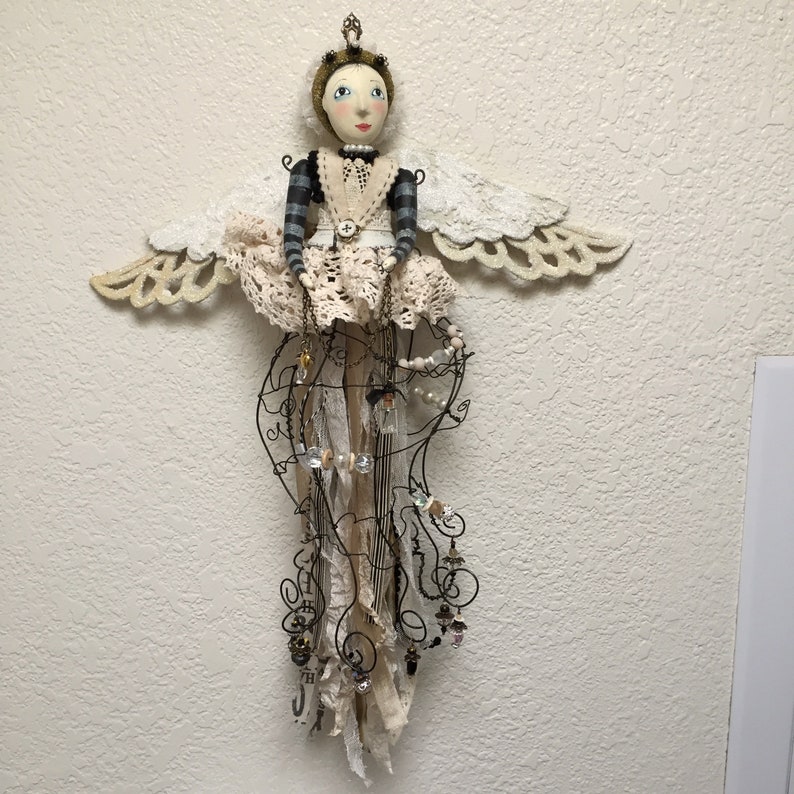 Pu&ograve; includere: Una bambola decorativa da parete con viso dipinto, corona dorata e ali in pizzo bianco. La bambola indossa una maglia a maniche lunghe a righe nere e grigie con un grembiule in pizzo bianco e una gonna fatta di fili, strisce di tessuto e perline.