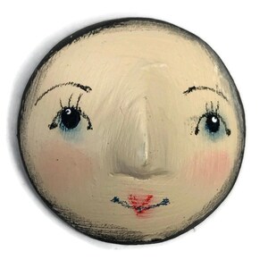 A flat-back round plaster doll head with hand-painted facial details including arching eyebrows, dark blue eyes, pink blush, and red lips.