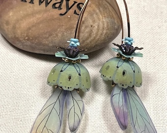 May include: A pair of dangle earrings featuring gold-toned curved wires, light green bell-shaped caps, and translucent, wing-shaped pendants in shades of pale blue, purple, and yellow. The earrings are displayed in front of a smooth brown stone with the word Always.