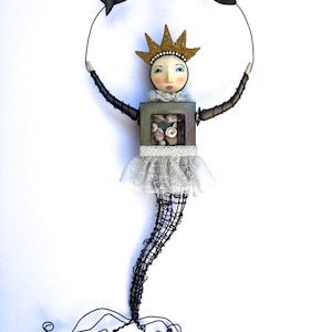 May include: A whimsical wire sculpture of a mermaid with a crown, holding a circle of wire with three small fish. The mermaid is made of wire and has a wooden torso with a lace skirt. The fish are made of wire and have black and gold paint.