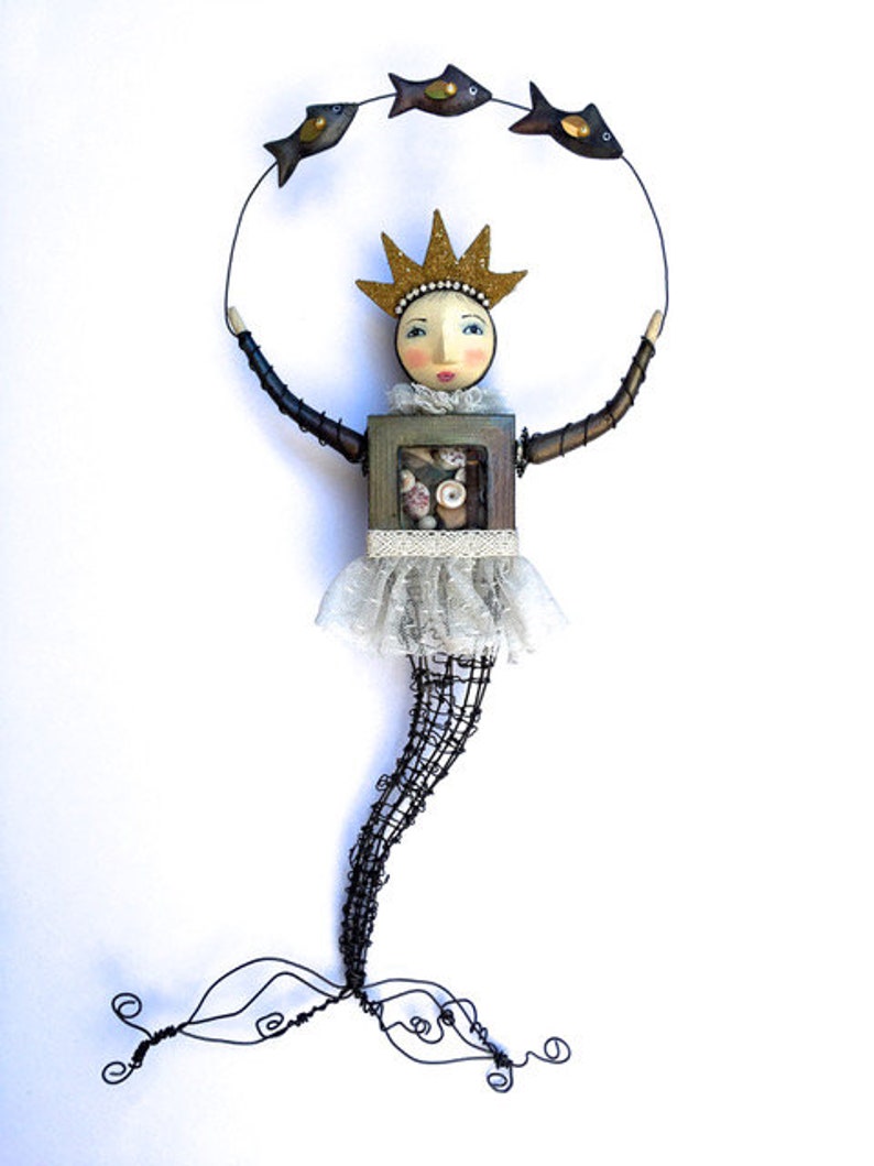 May include: A whimsical wire sculpture of a mermaid with a crown, holding a circle of wire with three small fish. The mermaid is made of wire and has a wooden torso with a lace skirt. The fish are made of wire and have black and gold paint.