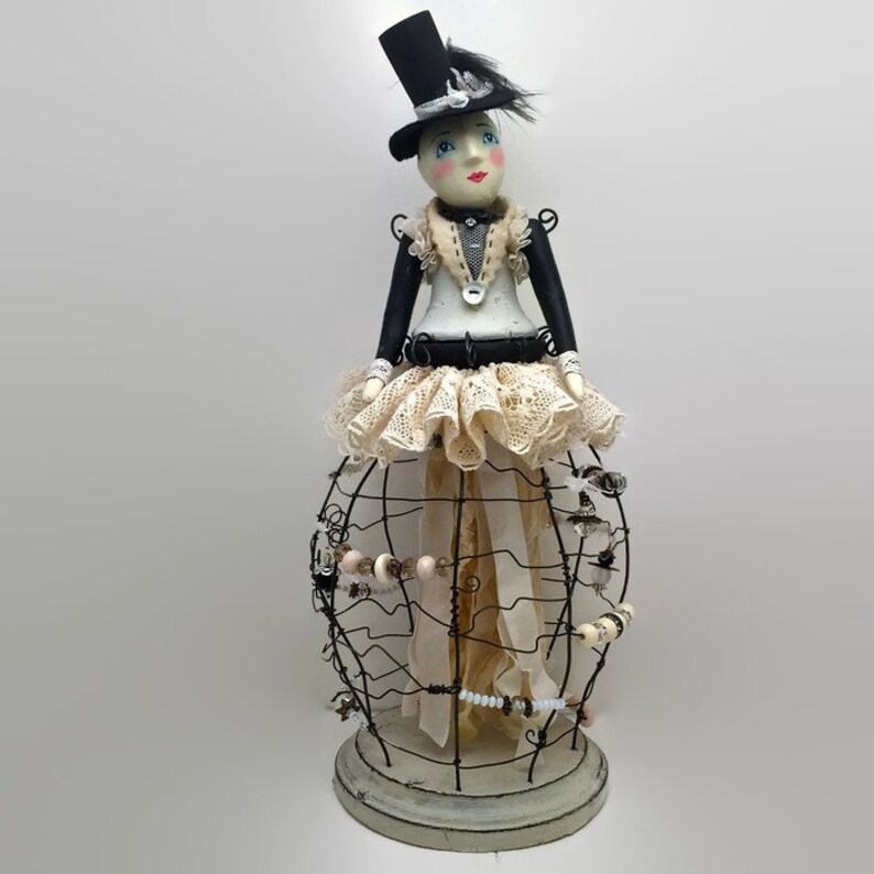 Pu&ograve; includere: Una bambola decorativa che indossa un cilindro nero con piume, un corpetto bianco e nero e una gonna in pizzo color crema. La parte inferiore della figura &egrave; racchiusa in una struttura a gabbia di filo metallico scuro decorata con perline.
