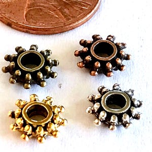 May include: Four small, round, gear-shaped metal beads with a central hole and radiating spikes along the outer edge. The beads are finished in antique bronze, copper, gold, and silver tones, shown next to a copper penny for scale reference.