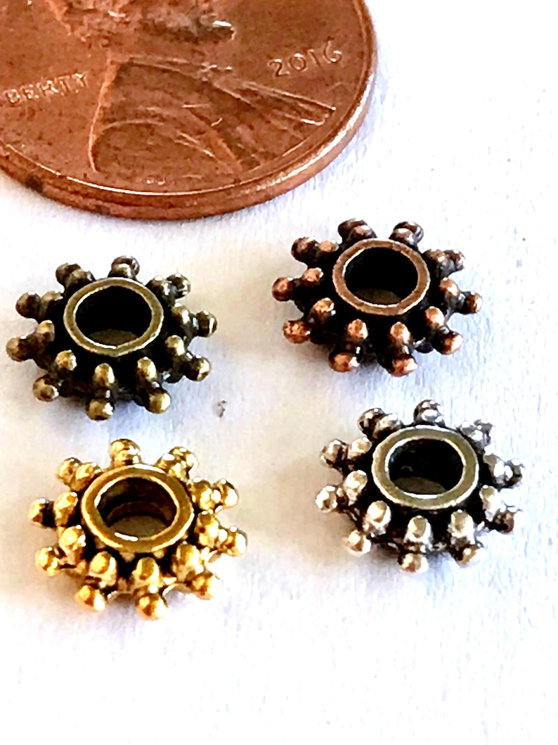May include: Four small, round, gear-shaped metal beads with a central hole and radiating spikes along the outer edge. The beads are finished in antique bronze, copper, gold, and silver tones, shown next to a copper penny for scale reference.