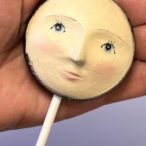 A hand holds a round plaster doll head on a stick that has been painted with facial features, blue eyes, rosy cheeks, and pink lips.