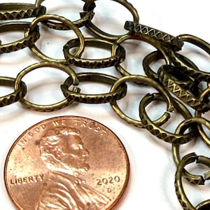 May include: A brass-toned metal chain with oval links featuring a textured, cross-hatched pattern on the surface. The chain is positioned next to a 2020 United States penny, which serves as a scale reference for the size of the individual chain links.