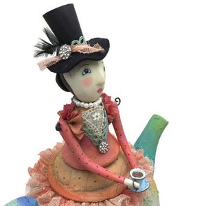 Pu&ograve; includere: Una statuetta decorativa di una persona che indossa un cilindro nero con una piuma e un dettaglio in strass, una collana di perle e un corpetto rosa a pois con bordo in pizzo, posizionata sopra una teiera dipinta con una gonna in tessuto color pesca.