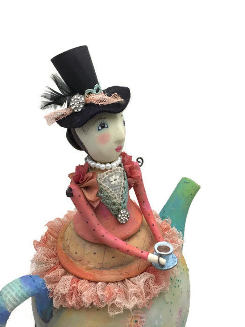 Pu&ograve; includere: Una statuetta decorativa di una persona che indossa un cilindro nero con una piuma e un dettaglio in strass, una collana di perle e un corpetto rosa a pois con bordo in pizzo, posizionata sopra una teiera dipinta con una gonna in tessuto color pesca.