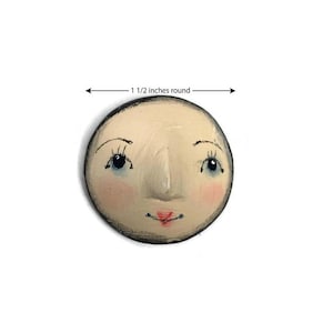 May include: A round, cream-colored face ornament with a painted expression featuring blue eyes, arched eyebrows, pink cheeks, and a small red mouth. The object has a dark outer edge and is labeled as measuring 1 1/2 inches round in the image.