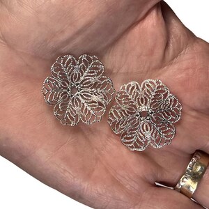 May include: Two silver-colored, filigree flower-shaped components. Each component has a detailed, openwork design with a central hole. The components are displayed on a light-colored background.