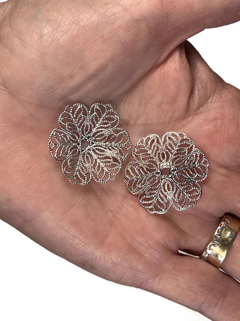 May include: Two silver-colored, filigree flower-shaped components. Each component has a detailed, openwork design with a central hole. The components are displayed on a light-colored background.