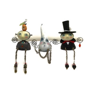Peut inclure: Trois figurines sur le th&egrave;me d'Halloween avec des visages peints et des jambes en fil de fer. La figurine de gauche a une citrouille sur la t&ecirc;te et un corps brun et rouge. La figurine du milieu est un fant&ocirc;me blanc avec une cha&icirc;ne autour du cou. La figurine de droite porte un chapeau haut de forme noir et un costume noir avec un n&oelig;ud papillon rouge.