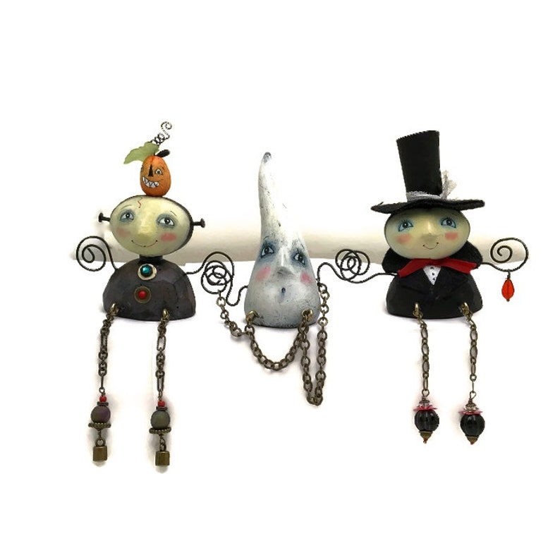 Peut inclure: Trois figurines sur le th&egrave;me d'Halloween avec des visages peints et des jambes en fil de fer. La figurine de gauche a une citrouille sur la t&ecirc;te et un corps brun et rouge. La figurine du milieu est un fant&ocirc;me blanc avec une cha&icirc;ne autour du cou. La figurine de droite porte un chapeau haut de forme noir et un costume noir avec un n&oelig;ud papillon rouge.