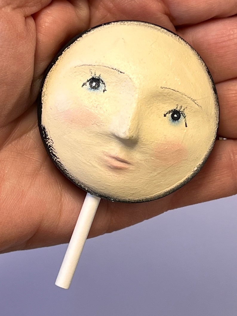 Puede incluir: Un objeto circular de color crema que presenta un rostro esculpido con ojos azules pintados, pesta&ntilde;as oscuras y mejillas de color rosa suave. La pieza tiene un borde exterior negro y un palo cil&iacute;ndrico blanco que sobresale de la parte inferior, similar a una piruleta.