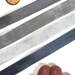 May include: Four strips of fabric trim in varying shades of grey and white, arranged diagonally. A copper-coloured US penny is positioned in the top left corner for scale, showing the width of the textured, woven material in this collection.