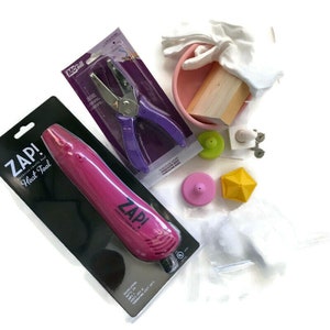 Könnte beinhalten: Ein rosa Heißluftwerkzeug mit der Aufschrift "Zap!" in weißer und schwarzer Verpackung. Das Werkzeug ist von Bastelutensilien umgeben, darunter ein violetter Locher, eine rosa Schüssel, ein Holzblock und weiße Handschuhe.