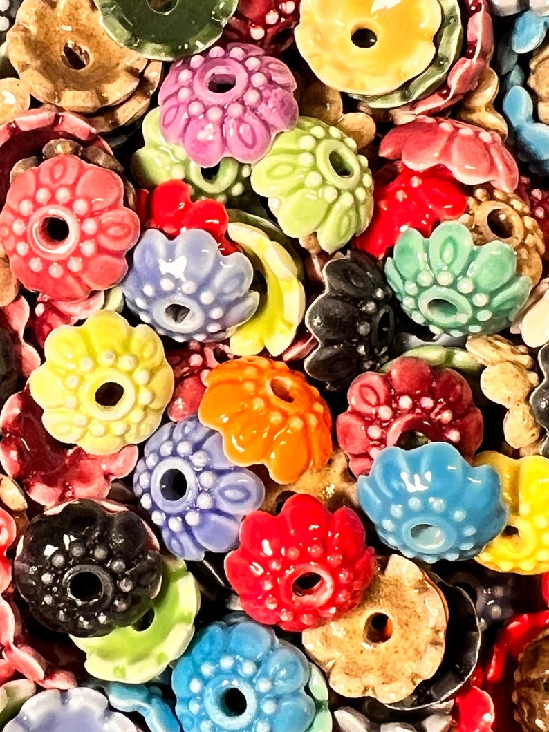 Pu&ograve; includere: Assortimento di perline di ceramica a forma di fiore colorate con un foro al centro. Le perline sono di vari colori, tra cui rosso, blu, verde, giallo, arancione e marrone.