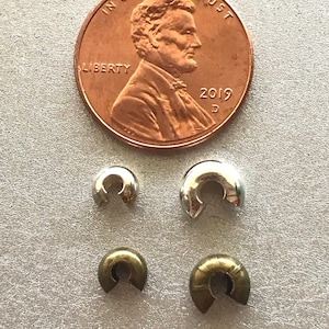 May include: A 2019 United States copper penny positioned above four small, C-shaped metal crimp covers. Two of the crimp covers are silver-toned and two are brass-toned, with one of each colour being slightly larger than the other.