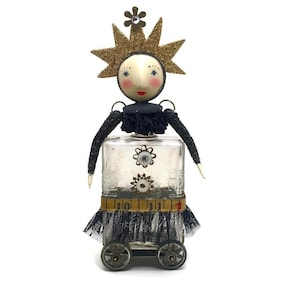 An art doll sculpture with a round plaster head, gold glitter star crown, glass bottle body with wheels, and dark fabric trim.