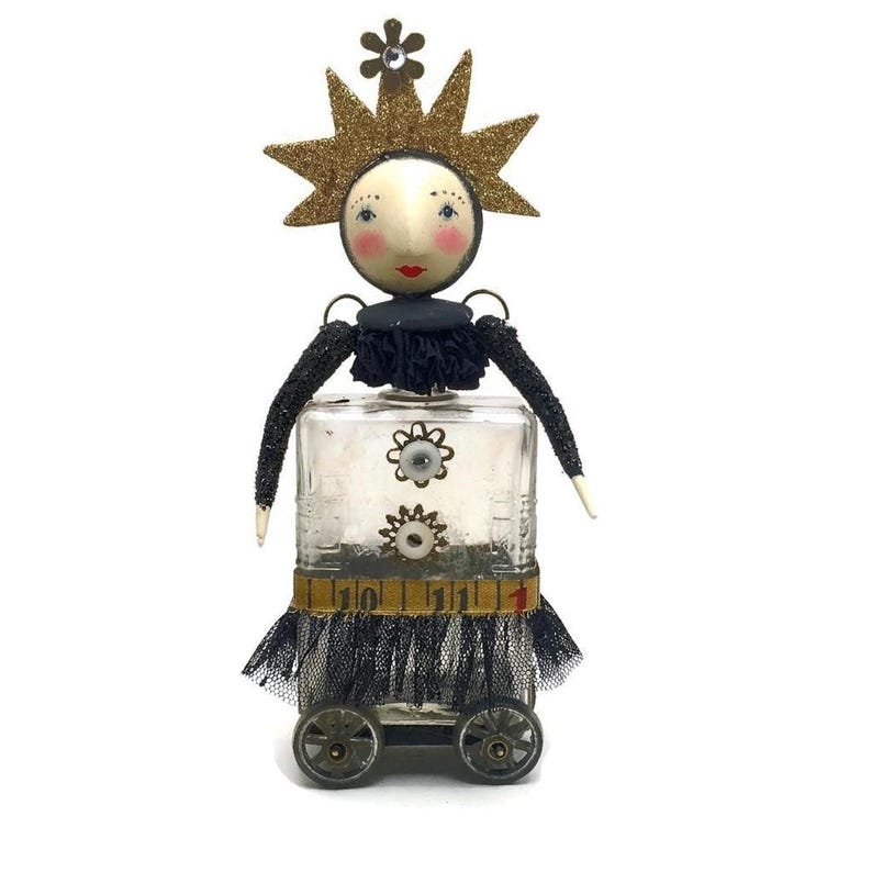 An art doll sculpture with a round plaster head, gold glitter star crown, glass bottle body with wheels, and dark fabric trim.