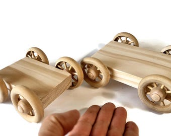May include: Two unfinished light-coloured wooden toy vehicles with rectangular platforms and four spoked wheels each. The wooden carts feature a natural grain texture and are constructed with simple, sturdy axles connecting the wheels.