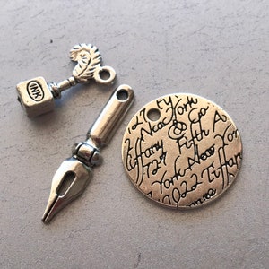 Könnte beinhalten: Drei silberne Anhänger: ein Schlüssel mit Federdesign, ein kleiner Würfel mit der Aufschrift "INK" und ein runder Anhänger mit der Aufschrift "Tiffany & Co. New York Fifth Ave. 10022 New York" in Kursivschrift.