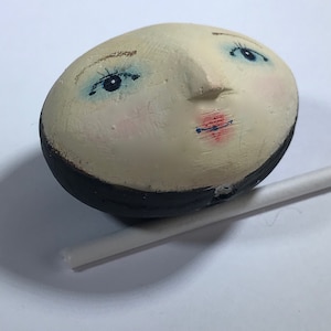 Peut inclure: Une t&ecirc;te de poup&eacute;e en c&eacute;ramique blanche avec un visage peint, des yeux bleus et un chapeau noir. La t&ecirc;te de poup&eacute;e est l&eacute;g&egrave;rement inclin&eacute;e sur le c&ocirc;t&eacute;.