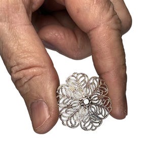 May include: A filigree flower-shaped jewelry piece with a silver-colored metal frame and white enamel filling. The delicate design features openwork petals and a central white accent. Held in a hand against a white background.