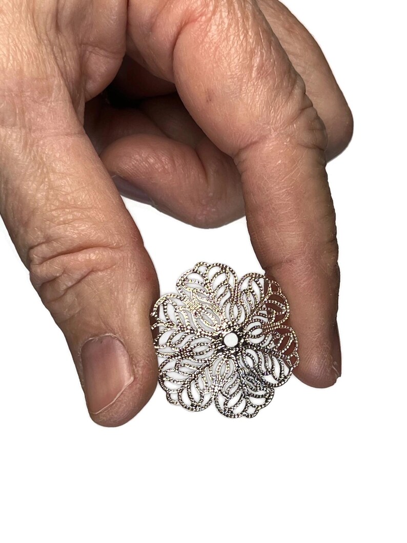May include: A filigree flower-shaped jewelry piece with a silver-colored metal frame and white enamel filling. The delicate design features openwork petals and a central white accent. Held in a hand against a white background.