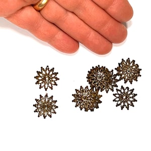 May include: A collection of five small, circular, antique bronze-colored metal filigree components. Each piece features a delicate, star-like or floral pattern with a central hole, designed with intricate cutouts and a textured, weathered metallic finish.
