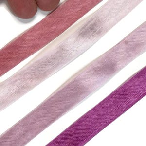 May include: Four strips of fabric bias tape arranged diagonally. The colors include a deep rose, a pale blush pink, a muted mauve, and a vibrant magenta. Each strip features a consistent woven texture and a folded edge along the length.