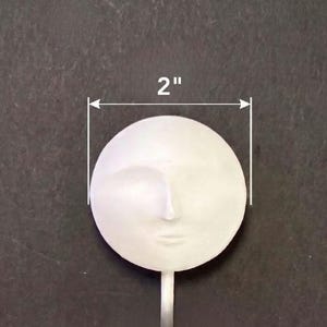 Puede incluir: Un objeto circular blanco que presenta un rostro humano esculpido con rasgos sutiles. El objeto mide 5,08 cm de di&aacute;metro y tiene aproximadamente 1,91 cm de profundidad, con un tallo delgado que se extiende desde la base del rostro.