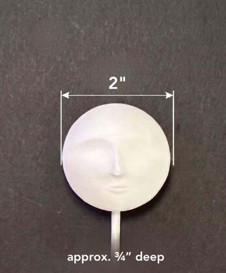 Puede incluir: Un objeto circular blanco que presenta un rostro humano esculpido con rasgos sutiles. El objeto mide 5,08 cm de di&aacute;metro y tiene aproximadamente 1,91 cm de profundidad, con un tallo delgado que se extiende desde la base del rostro.