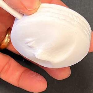 Puede incluir: Un objeto circular blanco de yeso o cer&aacute;mica con acabado mate que presenta un relieve de un rostro humano de perfil. El objeto tiene un borde liso y redondeado y una peque&ntilde;a varilla blanca unida a la parte superior que sobresale hacia afuera.