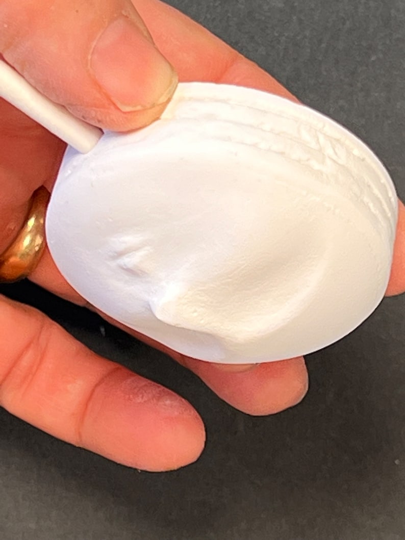Puede incluir: Un objeto circular blanco de yeso o cer&aacute;mica con acabado mate que presenta un relieve de un rostro humano de perfil. El objeto tiene un borde liso y redondeado y una peque&ntilde;a varilla blanca unida a la parte superior que sobresale hacia afuera.