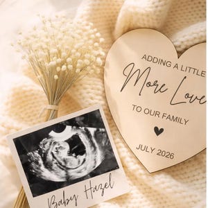 Peut inclure: Annonce de grossesse avec une photo d'échographie intitulée "Baby Hazel" et un cœur en bois avec le texte "Adding a Little More Love to Our Family" et "July 2026". Les éléments sont sur une couverture en tricot crème.