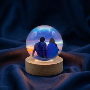 May include: A snow globe featuring a couple silhouetted against a sunset scene. The clear glass globe sits atop a wooden base that emits a warm glow. The background is a dark blue fabric.